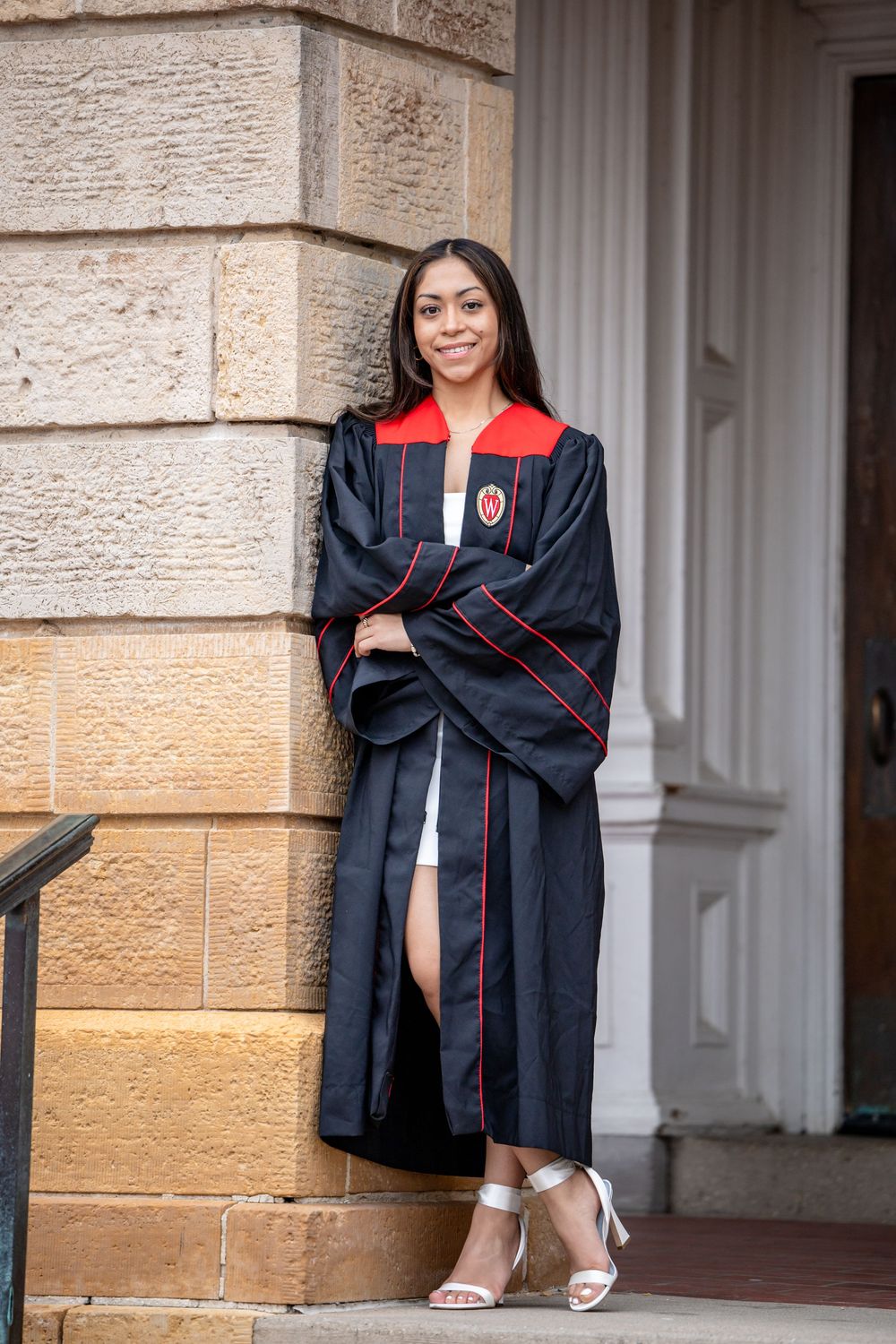 Graduate posing in academic regalia with black and red graduation gown on stone steps by white columns.