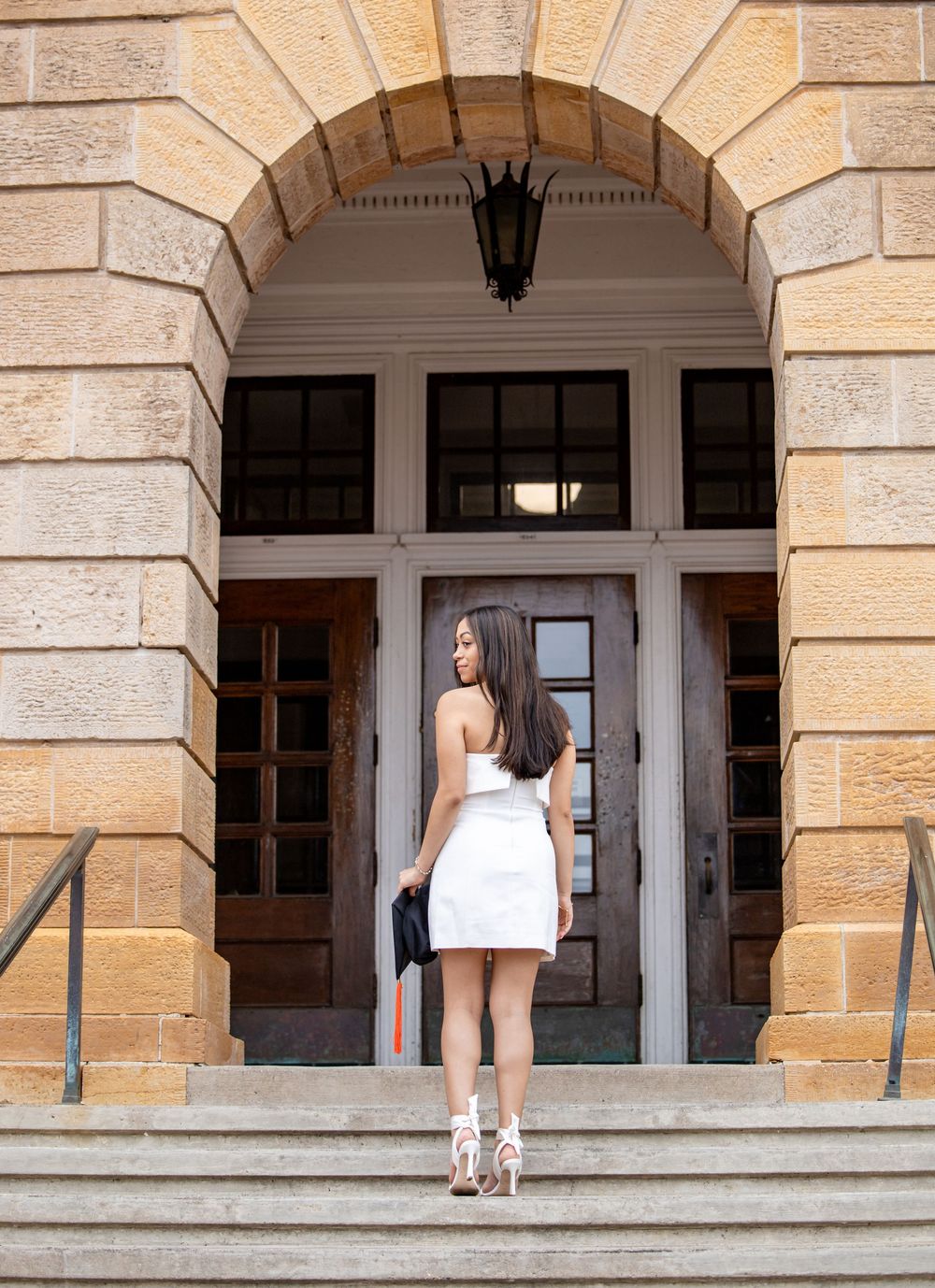 A person in a white dress and heels stands on stone steps before an arched entranceway with glass doors.