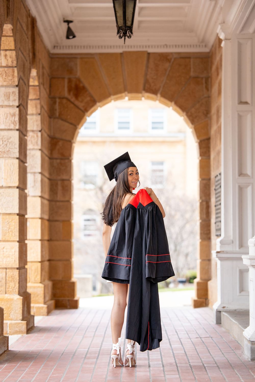 Graduate in black cap and gown poses in an elegant archway corridor while wearing stylish high heels.