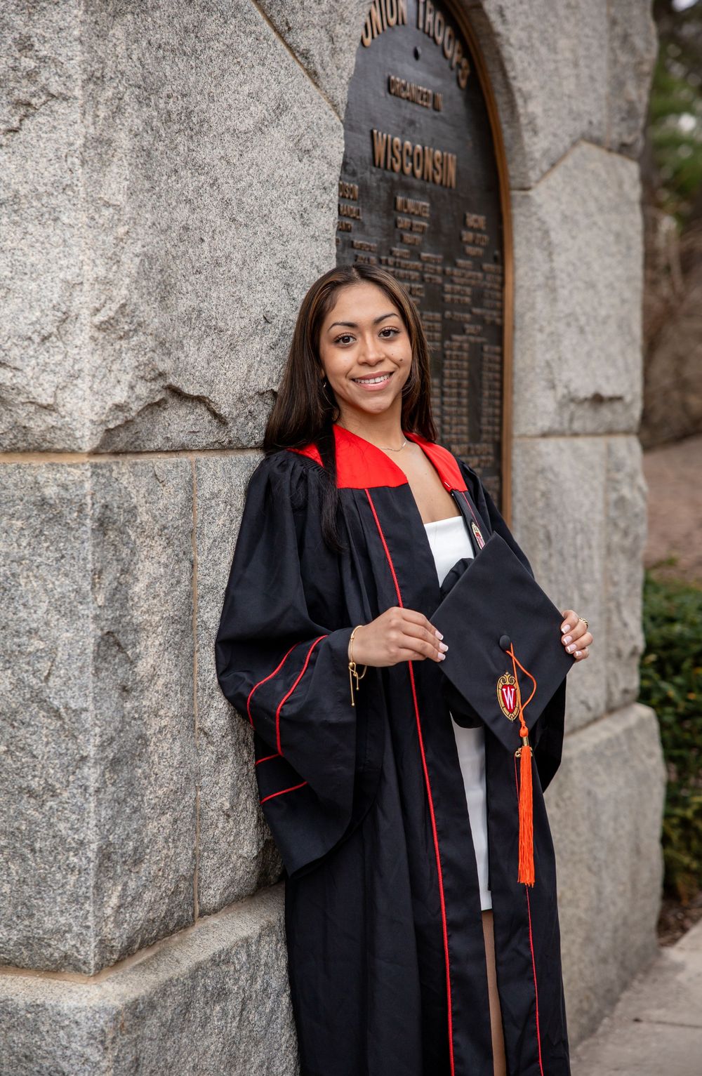 A graduate wearing a black academic gown with red trim stands smiling near a stone archway on graduation day.