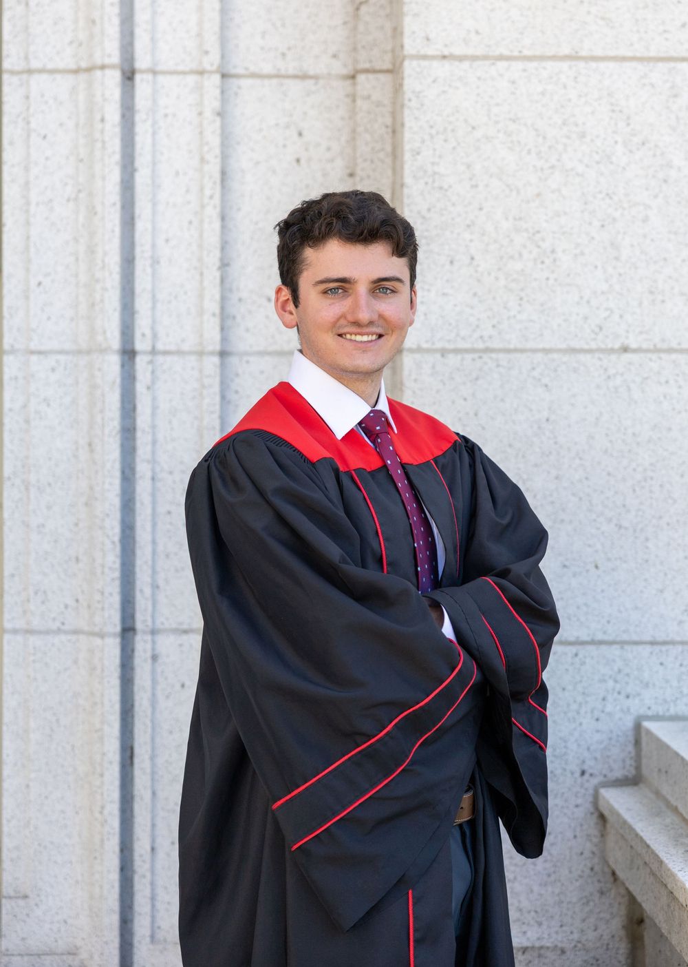 Graduate poses in traditional black academic regalia with red trim against stone wall background.