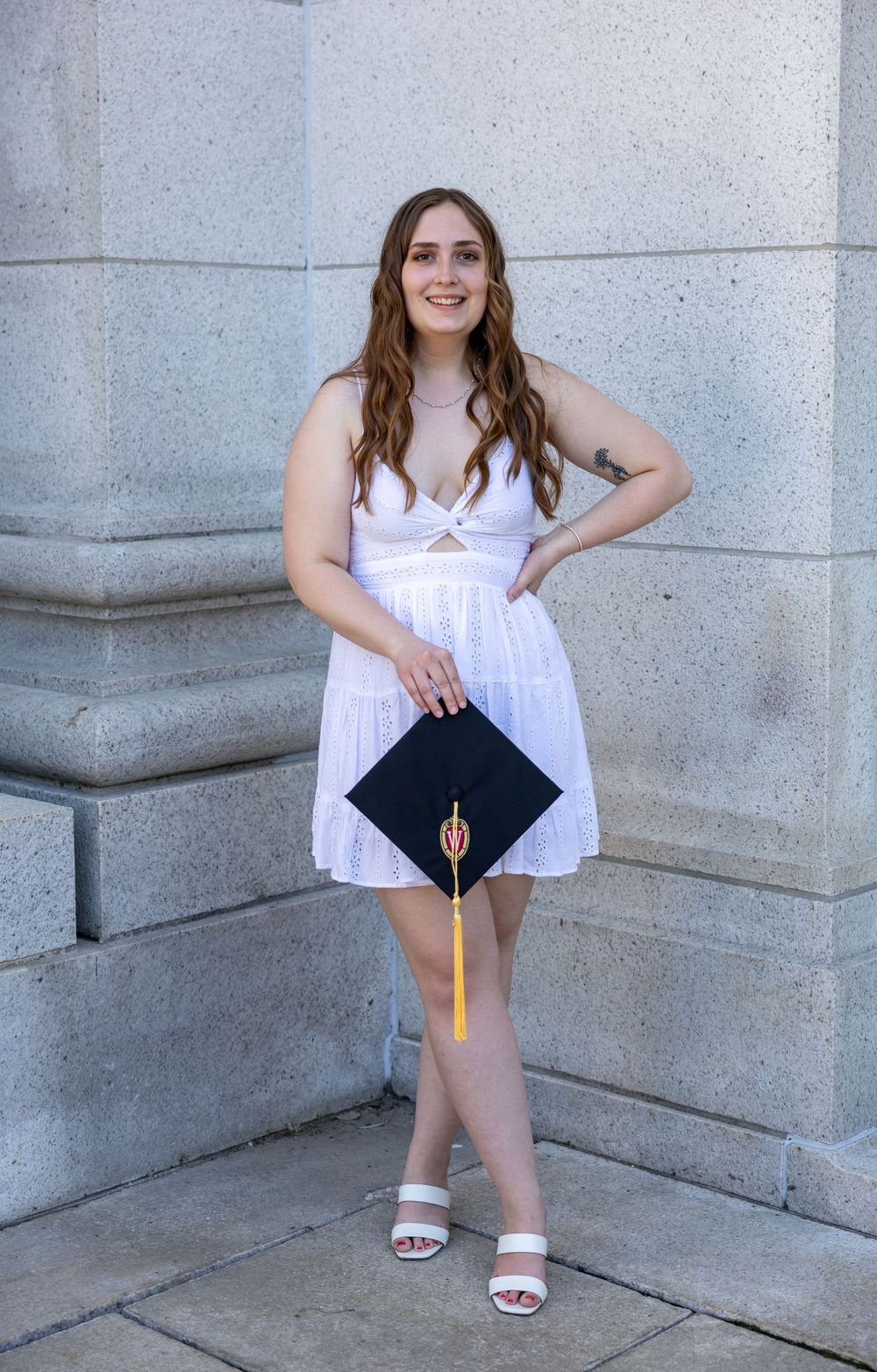 Graduate in a white summer dress holds diploma while posing against stone column architecture wearing sandals.