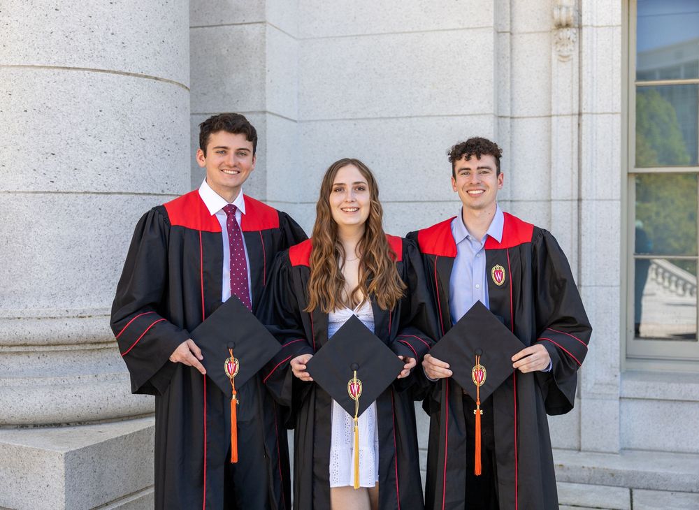 Graduates in red and black academic regalia pose together on stone steps of a university building.