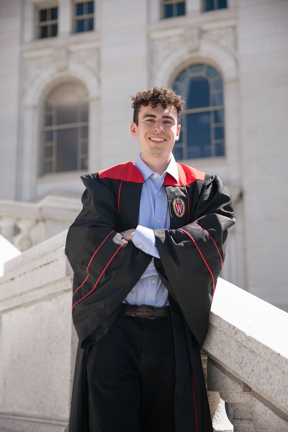 Graduate in black and red academic regalia stands smiling on white stone steps of stately building with arched windows.