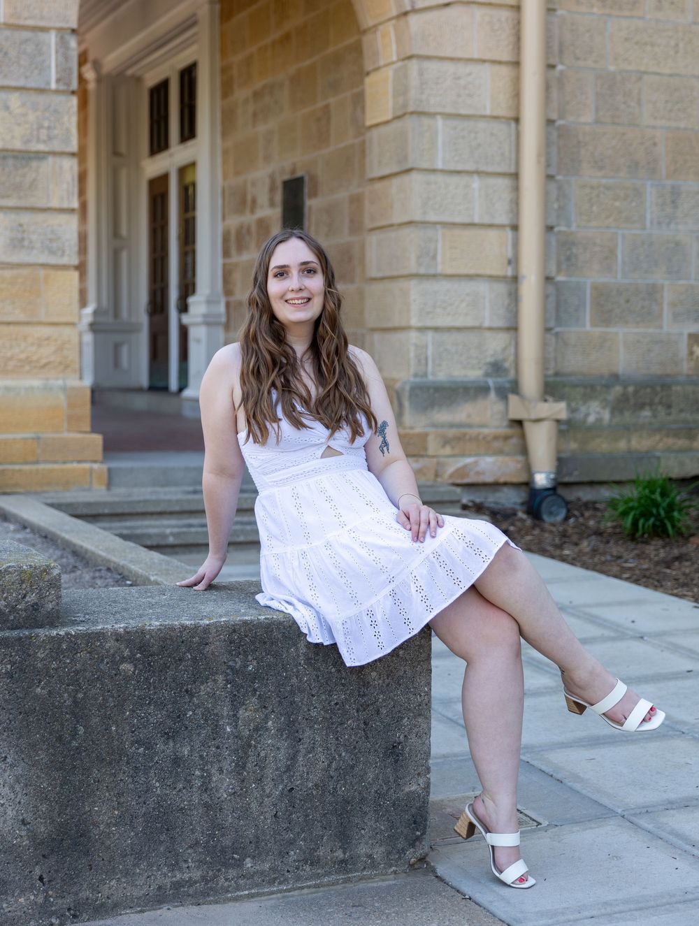 Someone wearing a white summer dress sits on a concrete ledge outside a historic stone building on a sunny day.