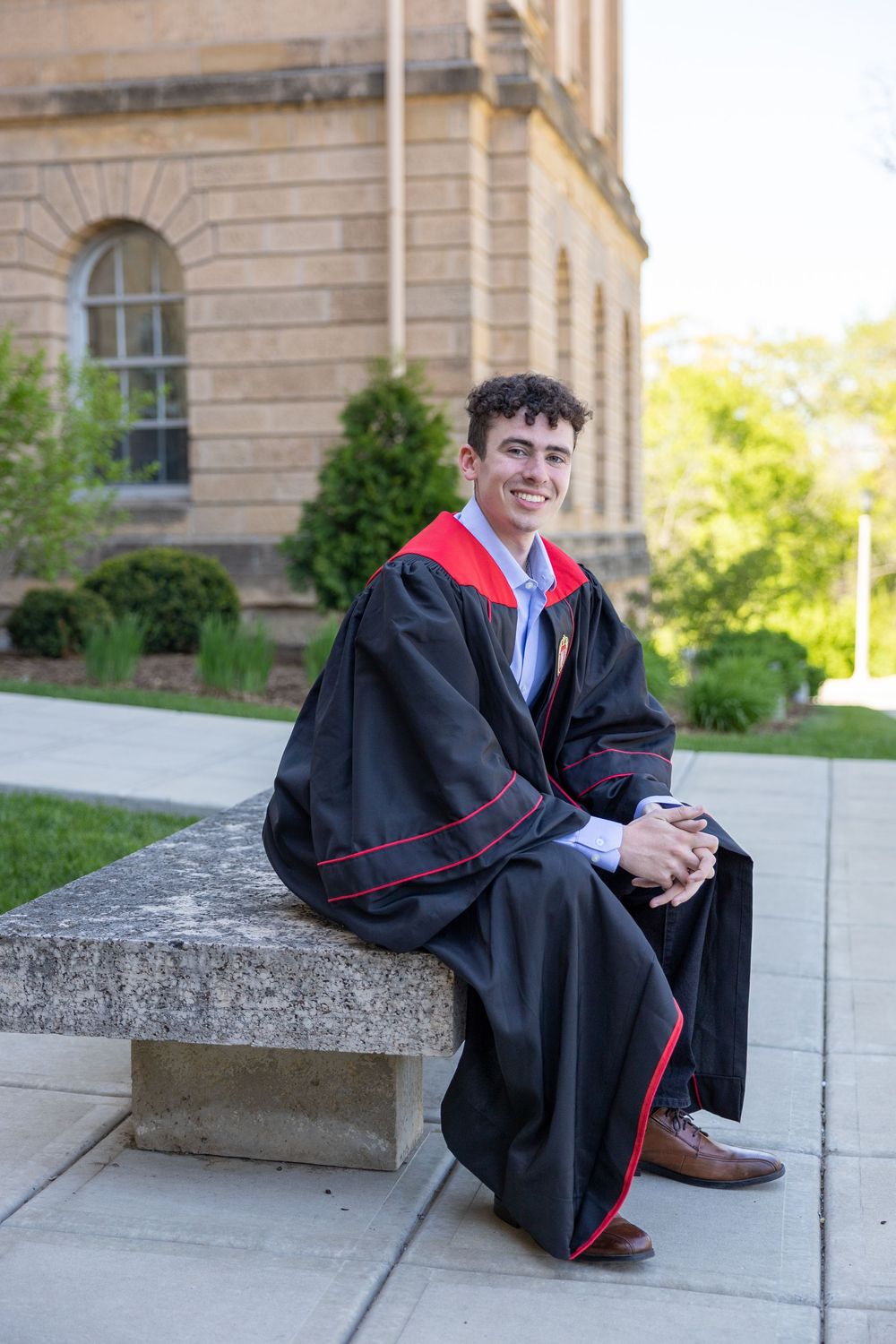 Graduate in black and red academic regalia sits smiling on stone steps outside a traditional brick building.
