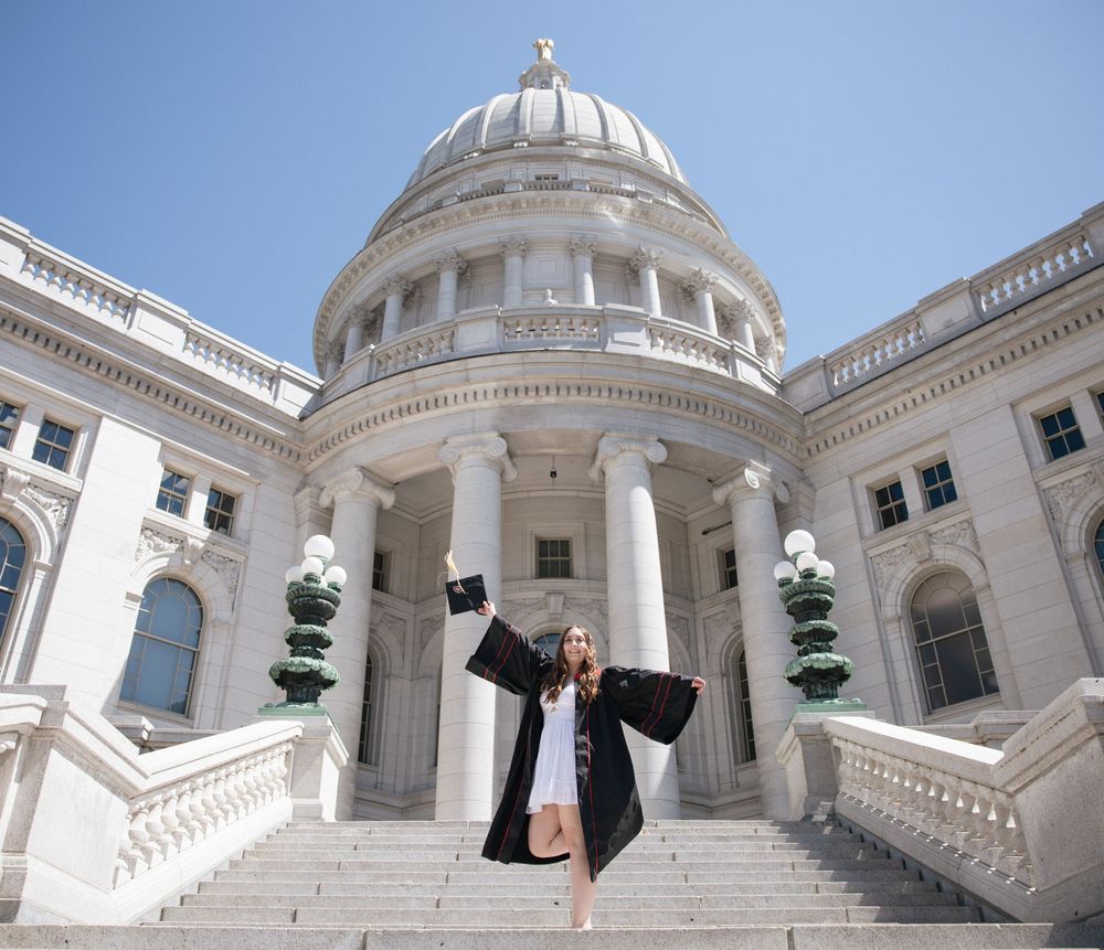 A graduate celebrates on the steps of the Wisconsin State Capitol building's grand entrance with its iconic white dome.