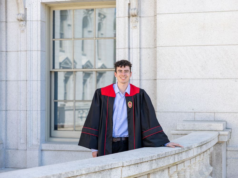 A graduate in academic regalia poses on a white stone balcony wearing a black graduation gown with red trim.