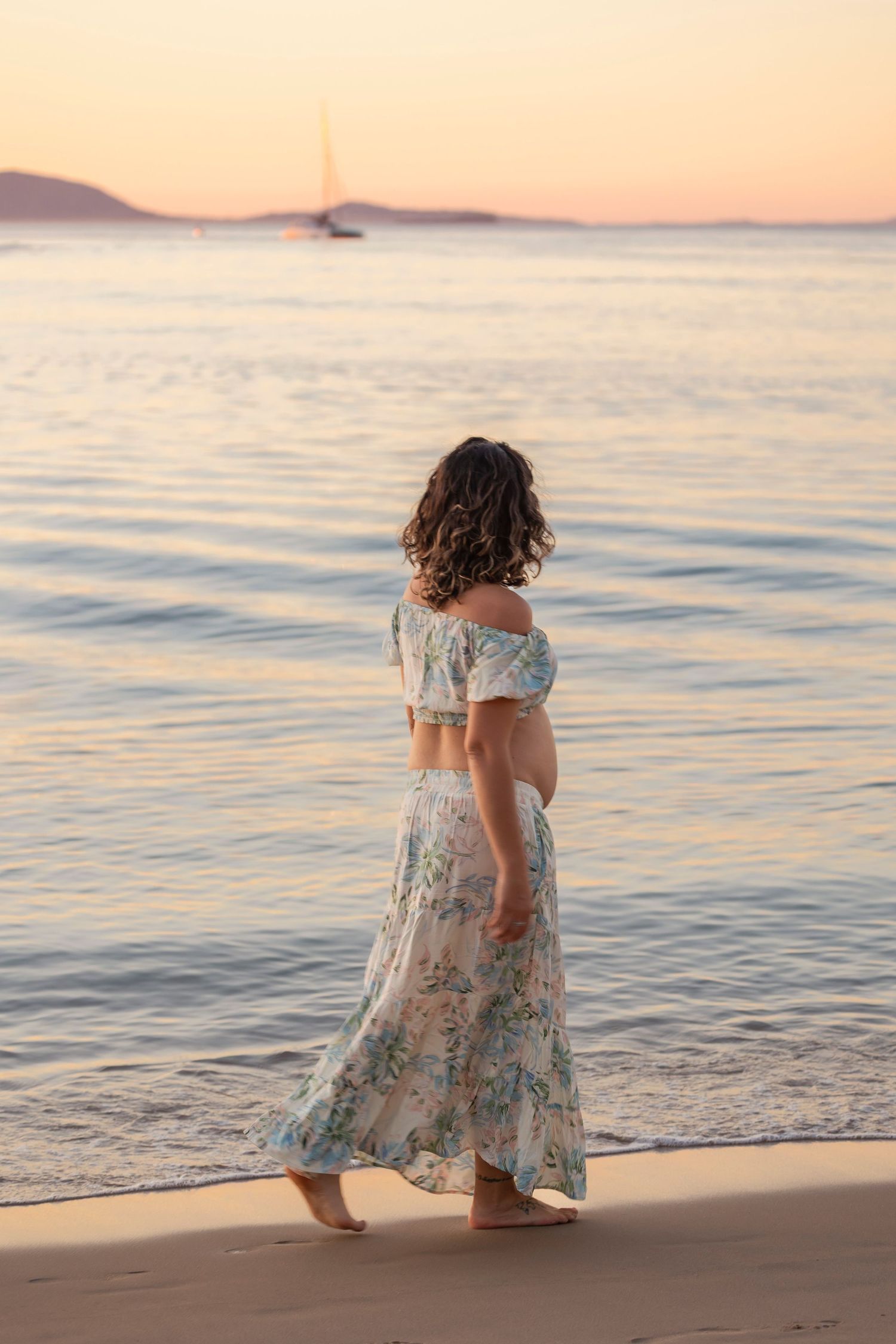 Person in flowing dress walking along beach at sunset.