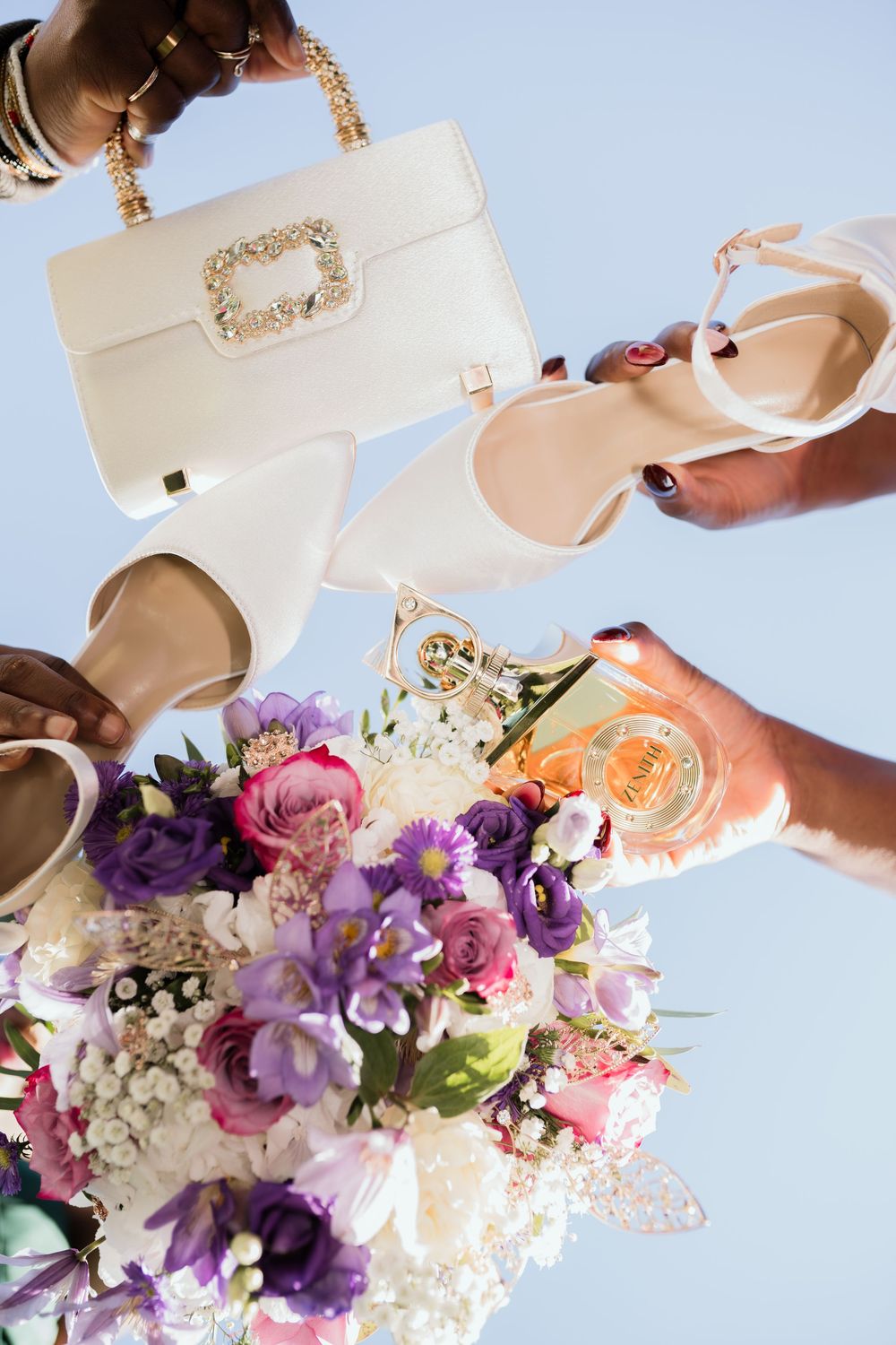 Bridal flat lay with white heels, pearl-embellished handbag, purple and pink flower bouquet, and gold jewelry against blue sky.