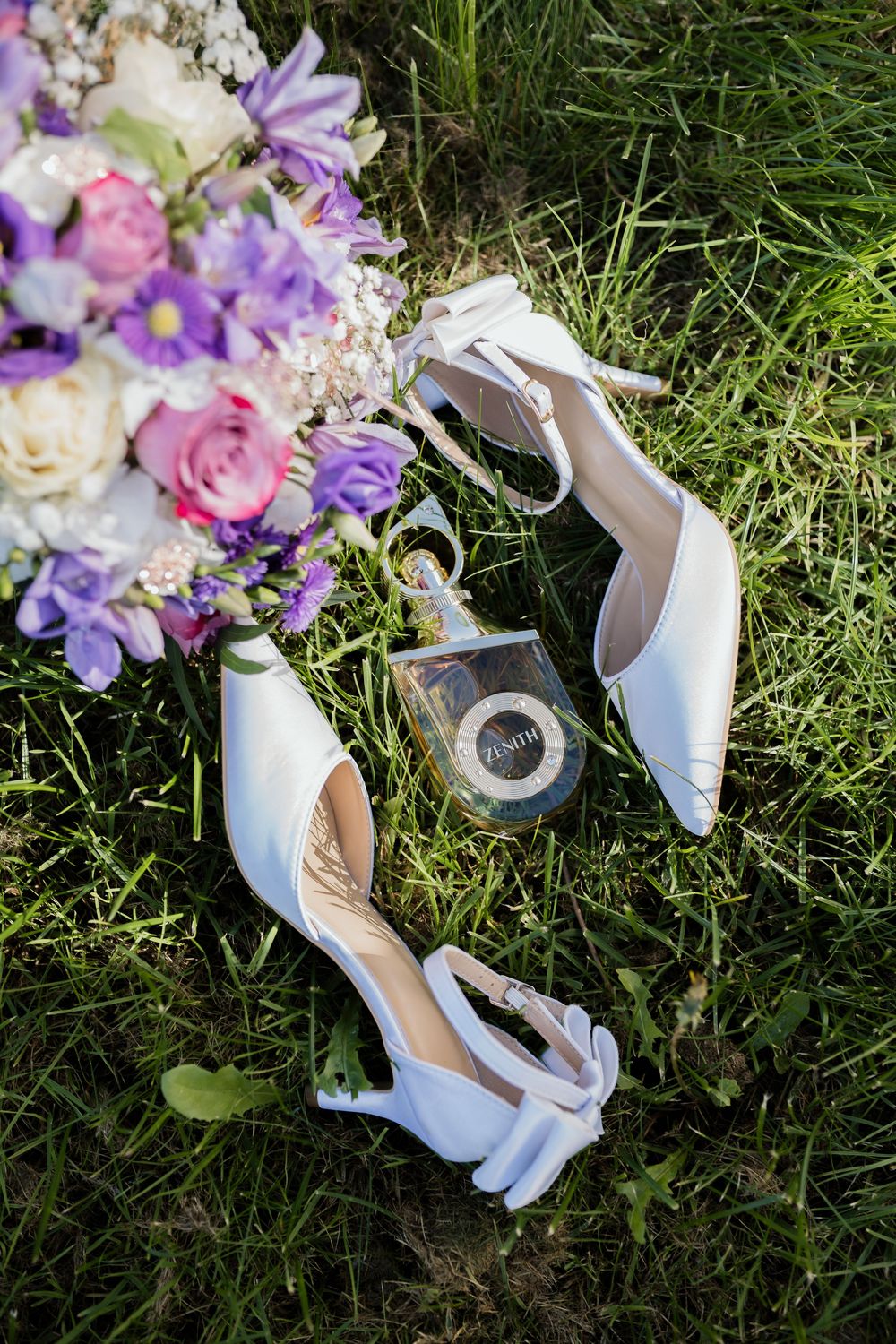 White bridal heels and perfume bottle resting on green grass beside a colorful wedding bouquet with purple and pink flowers.