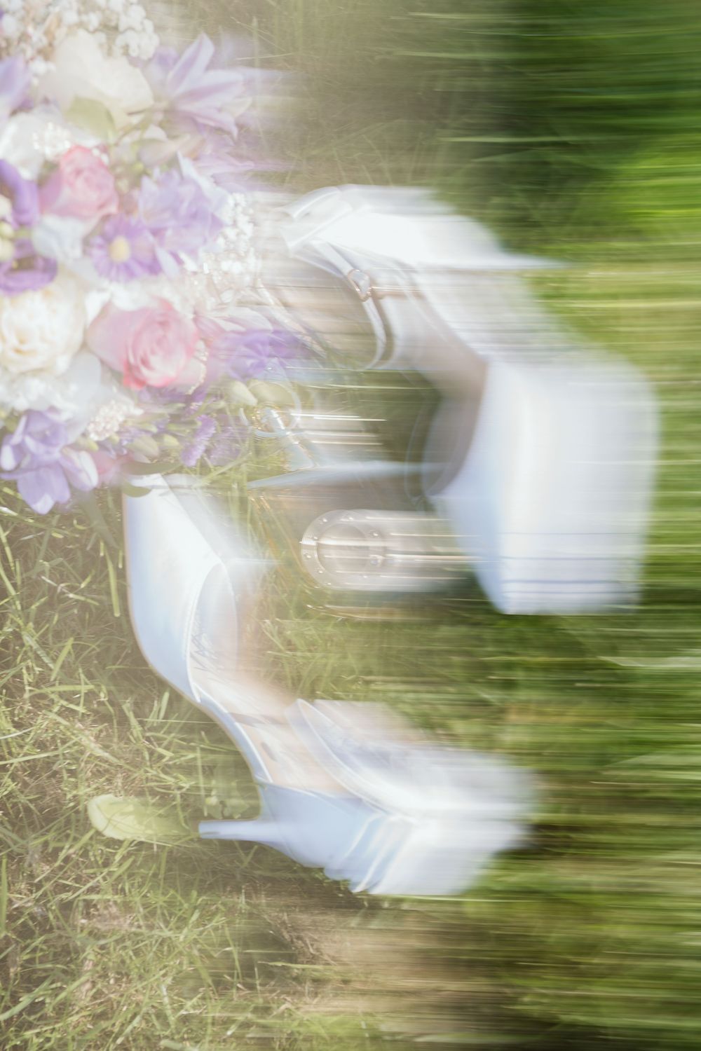 Blurred motion shot of white bridal heels beside a pastel floral bouquet on green grass.