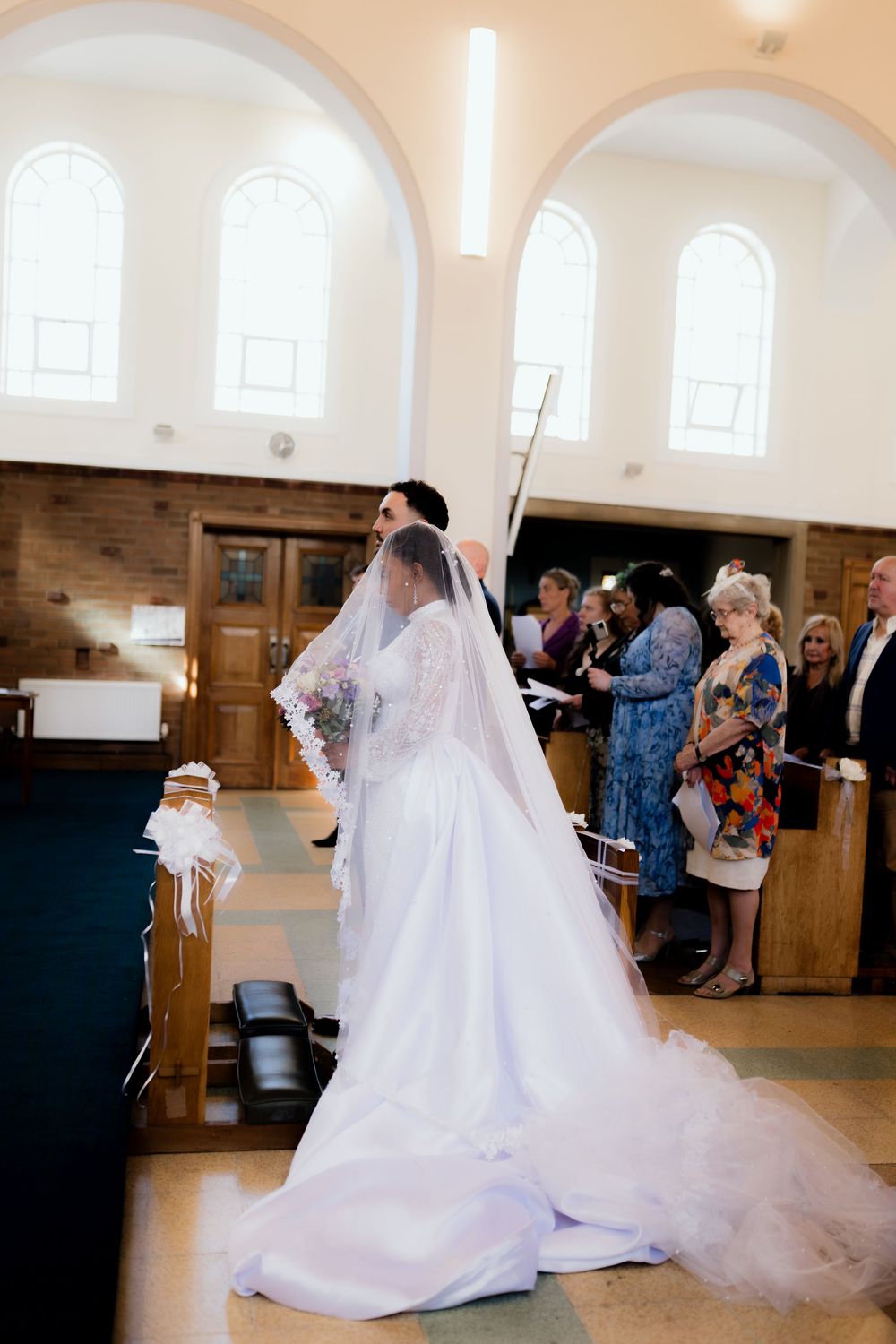 Bride in white gown and veil walks down church aisle with bouquet during wedding ceremony.