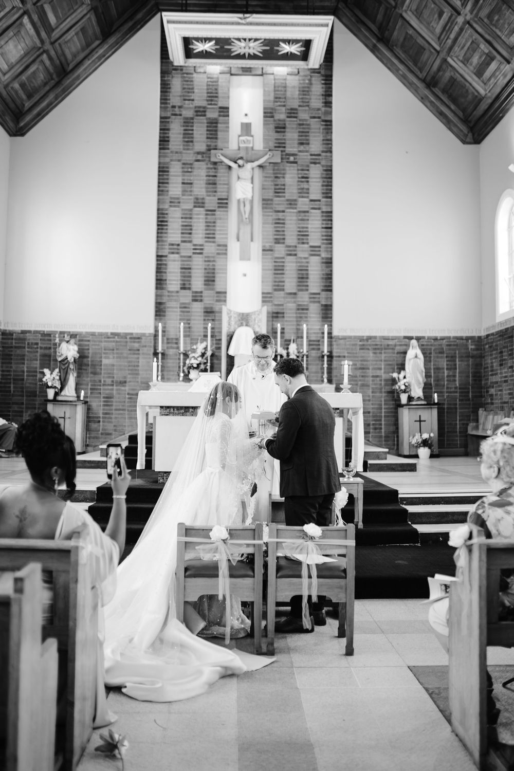 Bride and groom kneeling at altar during Catholic wedding ceremony in black and white church photo.