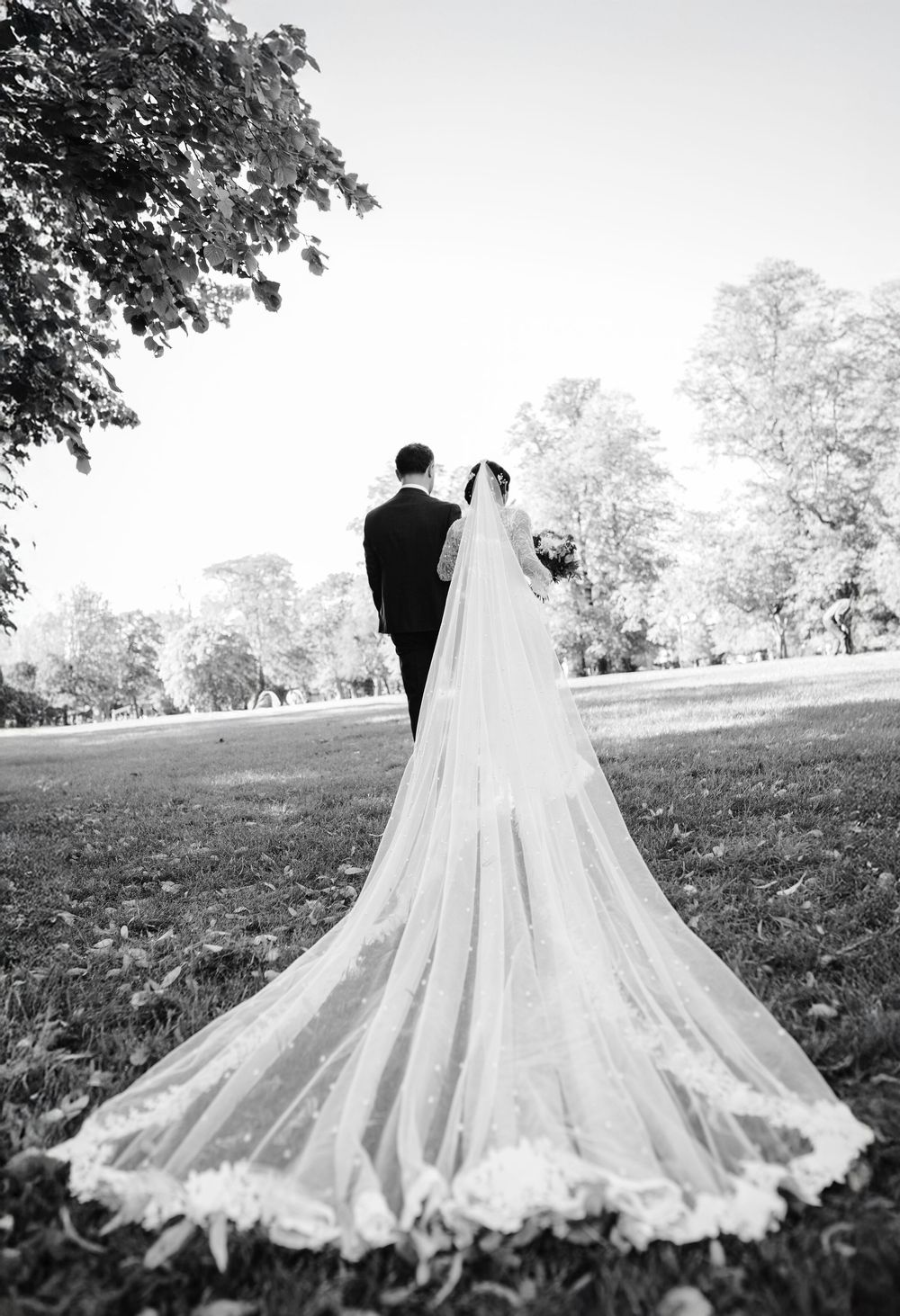 Black and white photo of a bride and groom walking in a park, her long lace cathedral veil trailing behind.