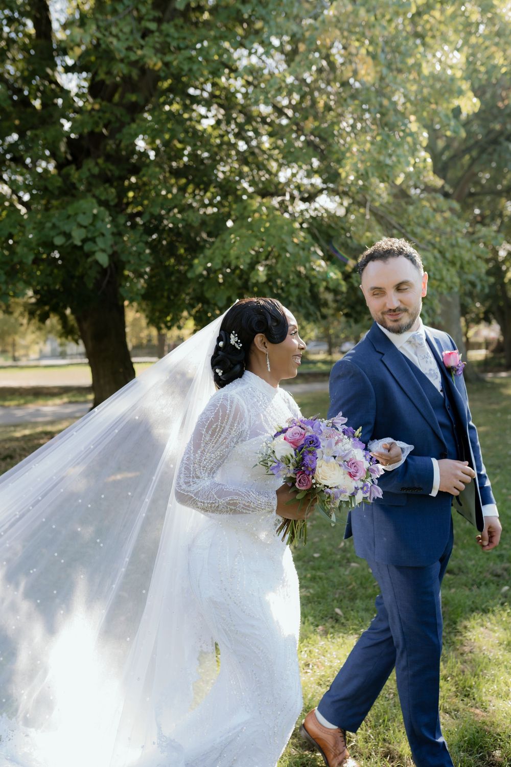 A bride and groom walk outdoors in a park, she in a white gown with veil, he in a navy suit holding flowers.
