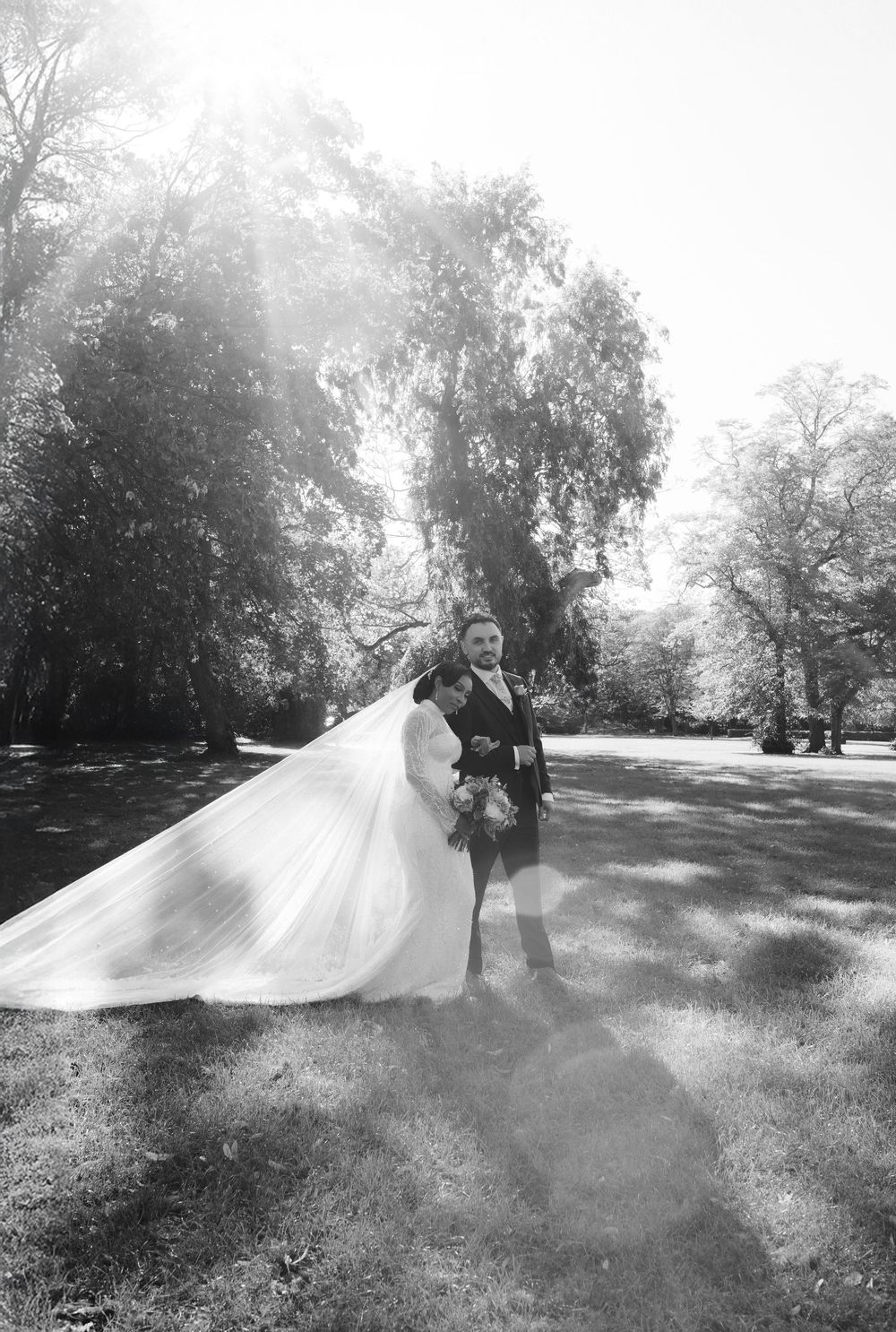 Black and white wedding photo of bride and groom embracing in sunlit park, veil flowing, groom in kilt.