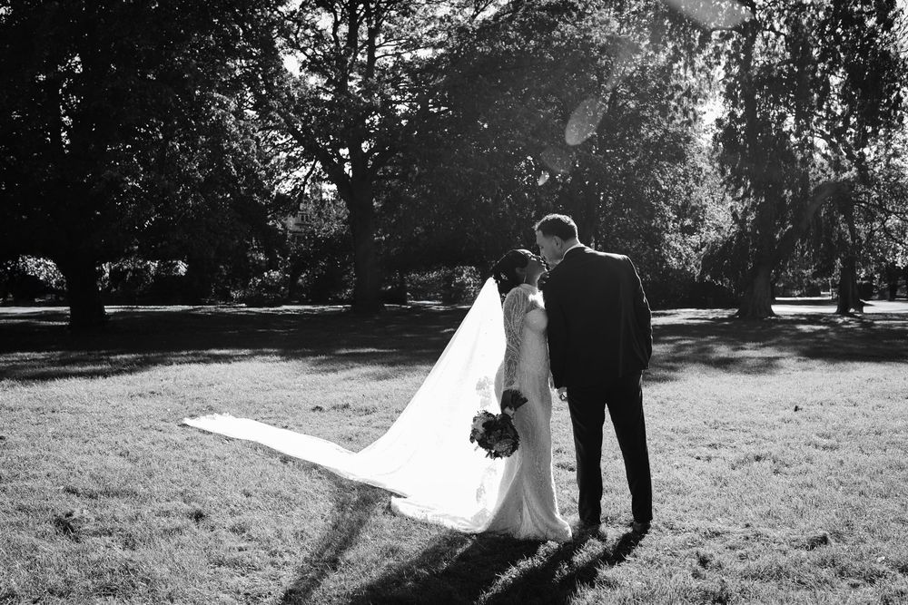 Black and white photo of a bride and groom standing together on a sunny lawn, with her veil trailing behind.