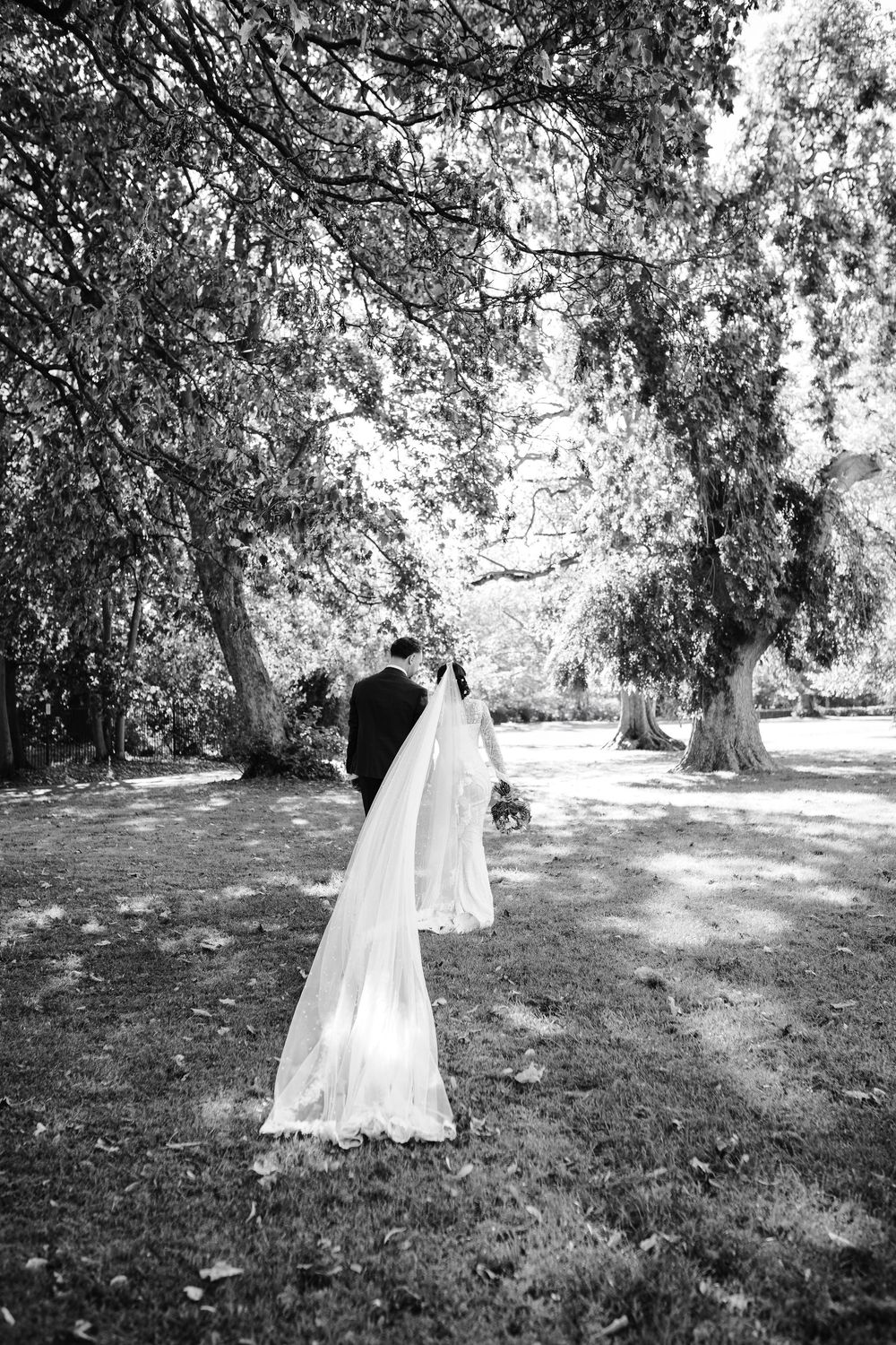 Black and white photo of bride and groom walking under large trees in park.
