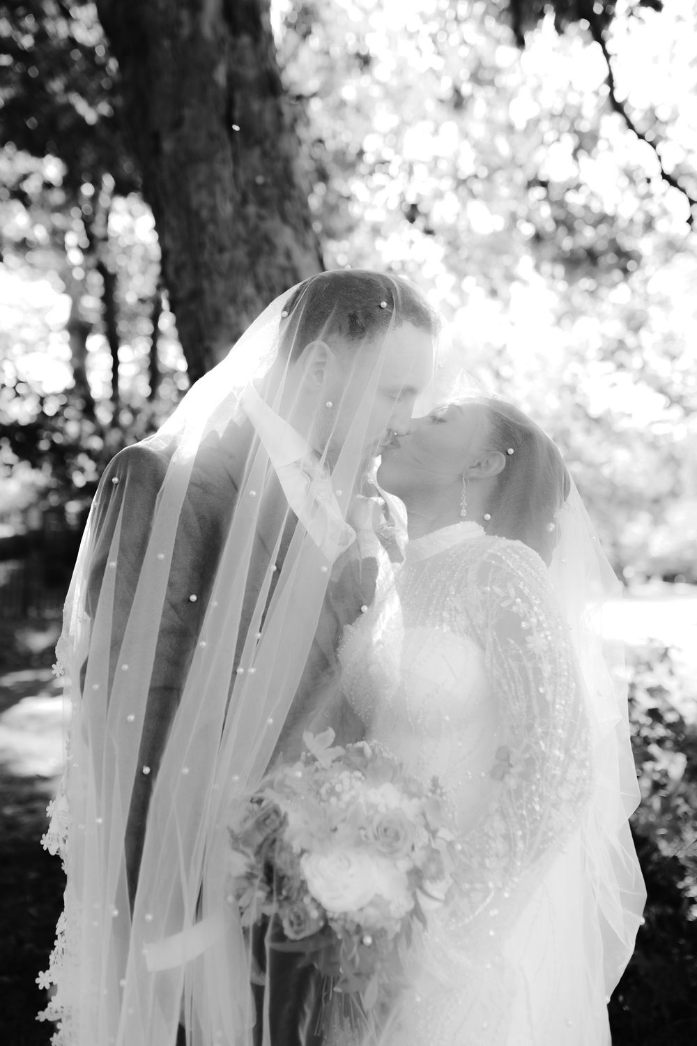 Black and white romantic photo of bride and groom kissing under flowing veil beside a tree.