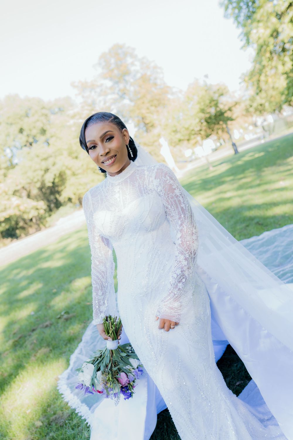 Radiant Black bride in long-sleeve lace wedding gown with cathedral veil holds purple bouquet outdoors.