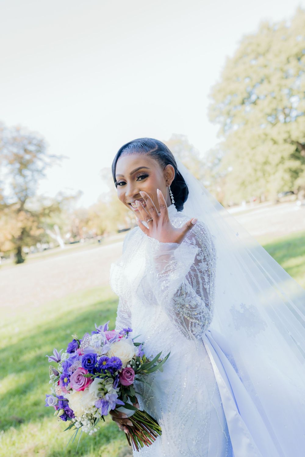 Smiling bride in white lace gown showing ring, holding purple bouquet outdoors.