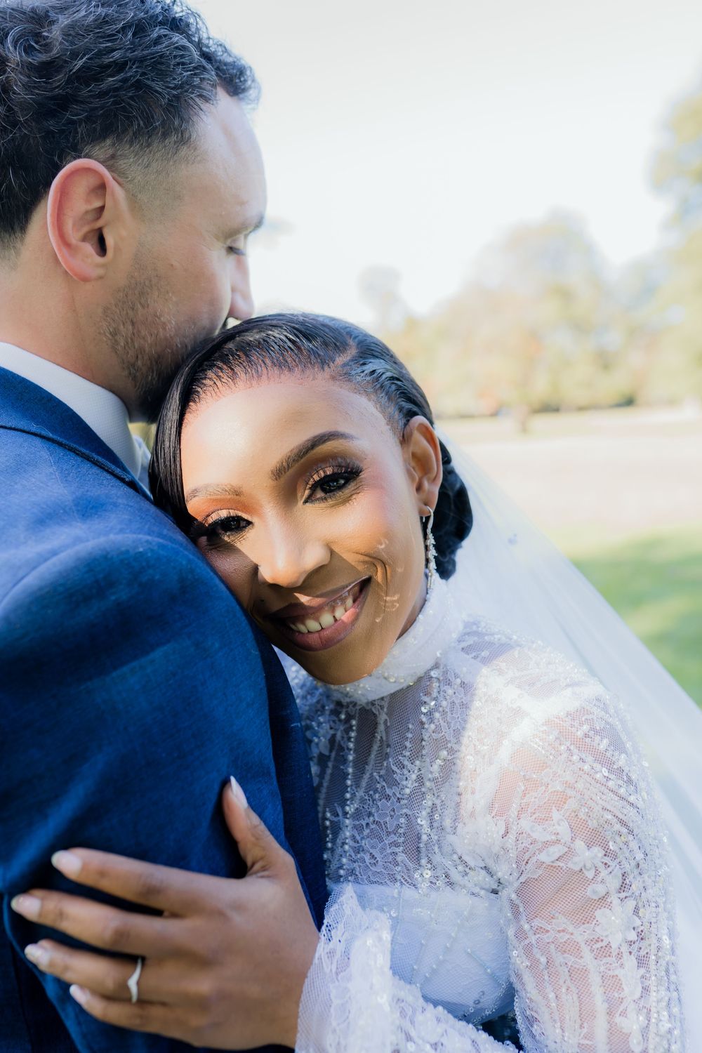 Bride in lace gown smiles while groom in blue suit kisses her cheek outdoors at a wedding ceremony.