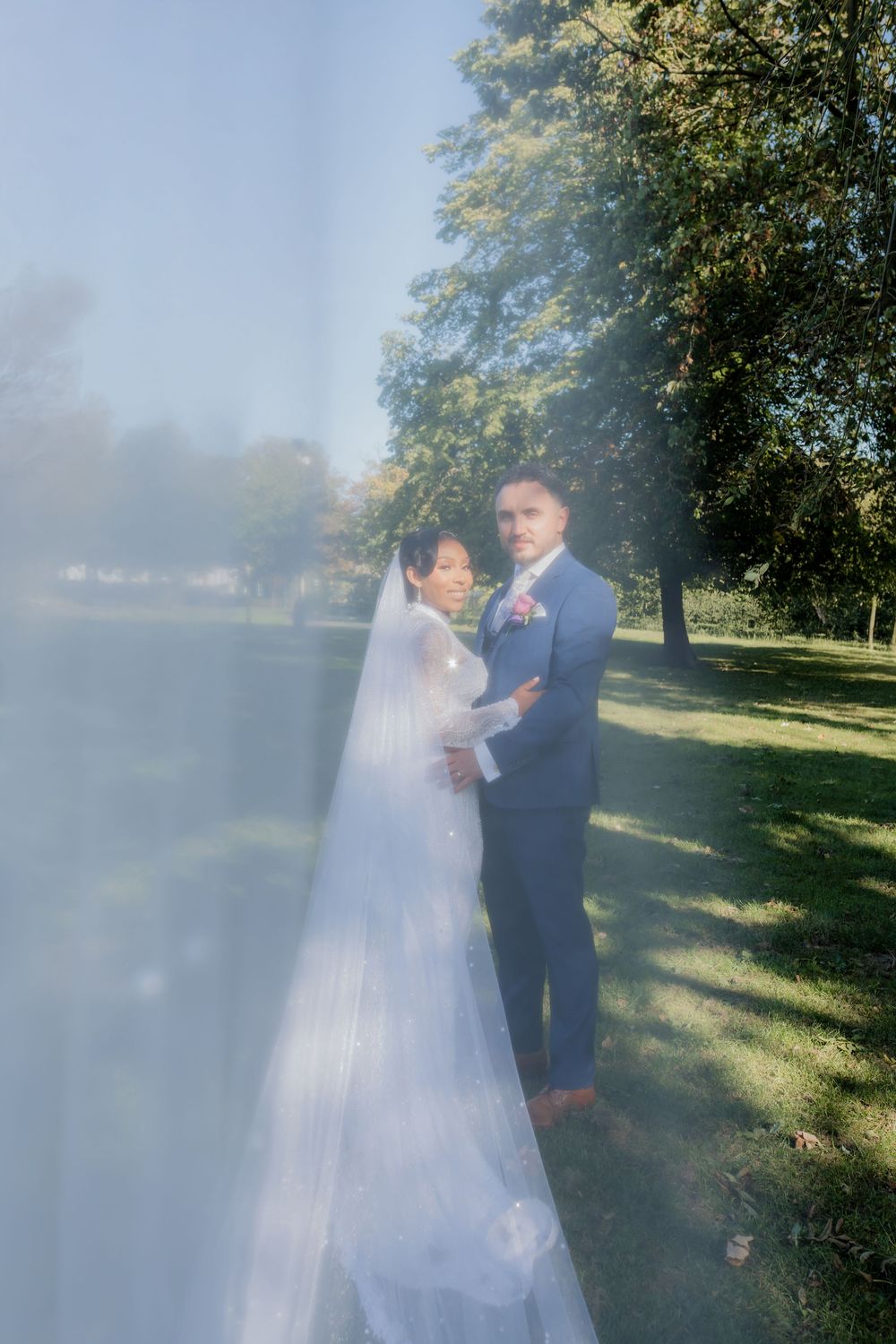 Bride in white gown and veil stands with groom in blue suit outdoors in a sunlit park on their wedding day.