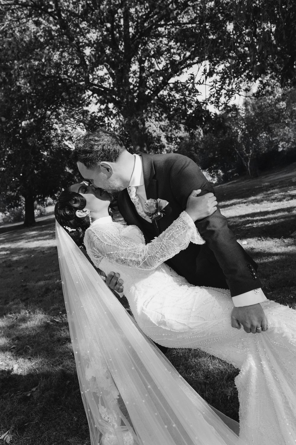 Black and white romantic dip kiss between bride in lace gown and groom under large park trees.