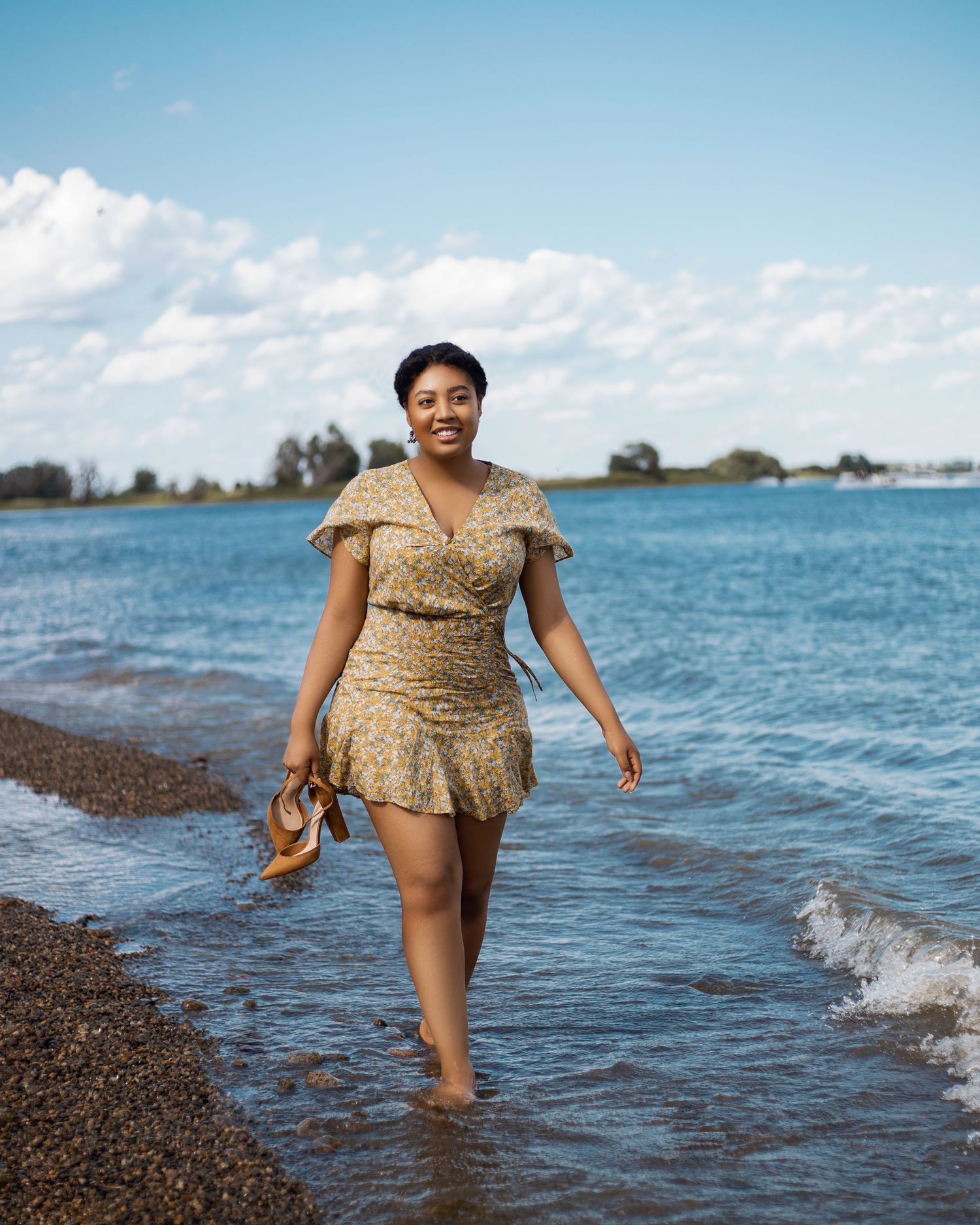 Woman in floral dress walks along beach shoreline holding sandals.