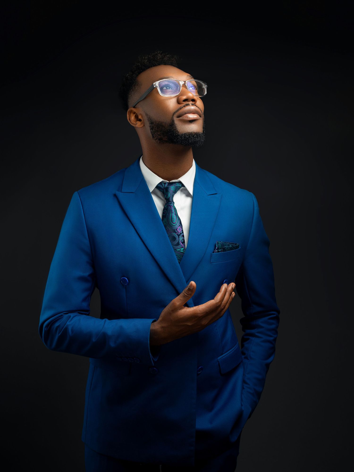 Professional portrait of person in blue suit and glasses posing confidently against dark background.