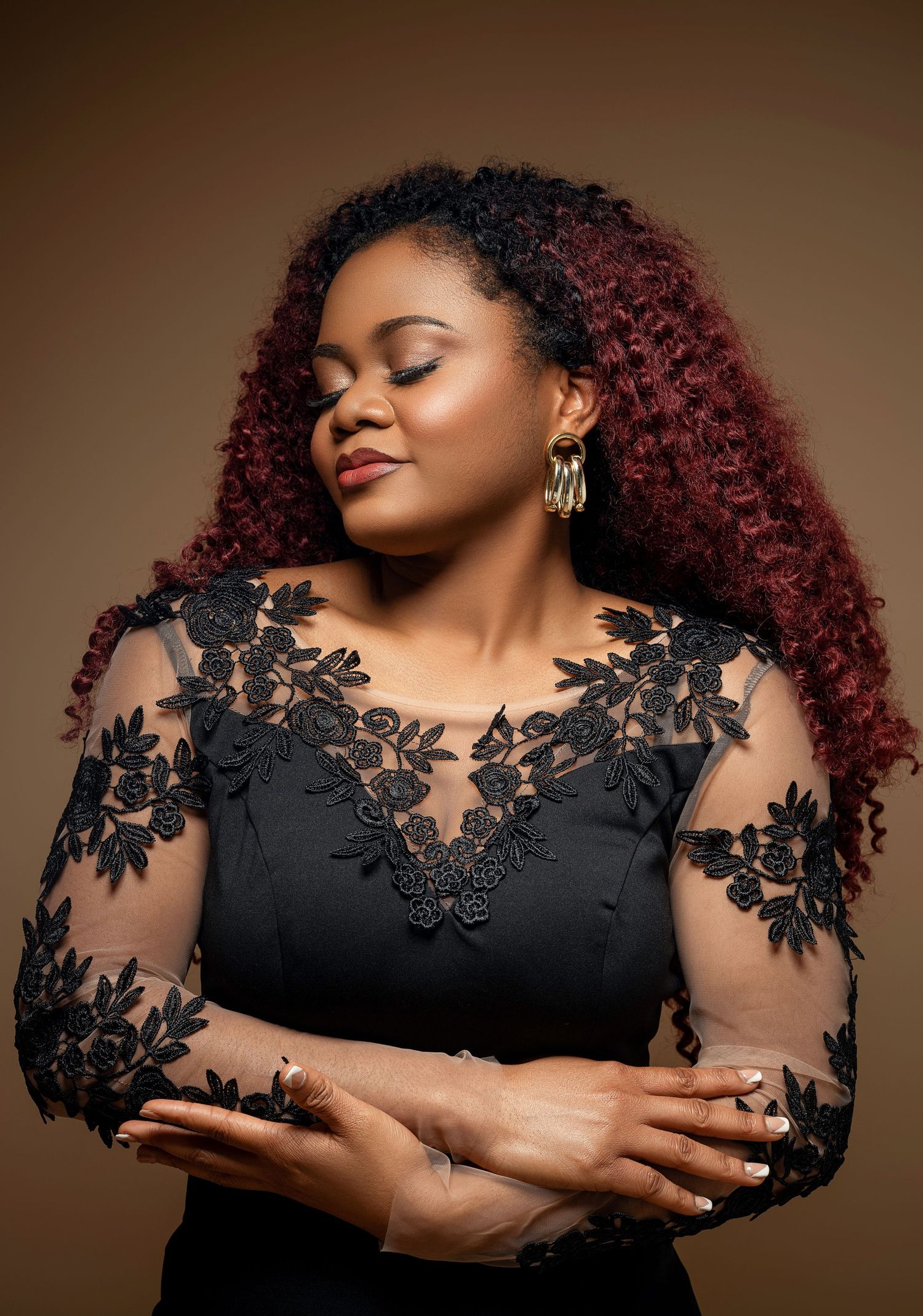 Woman with burgundy curls wearing elegant black lace dress poses against brown background.