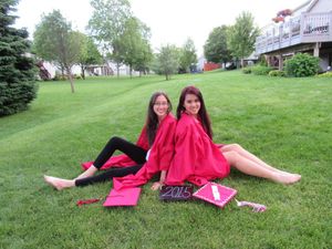 Two graduates in red attire relaxing on a manicured lawn with pink graduation materials.
