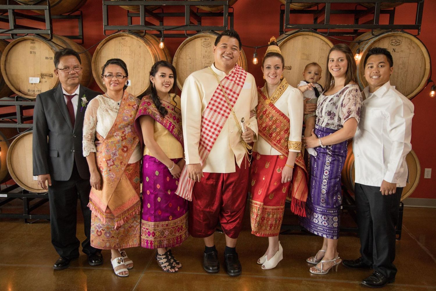 Group photo of wedding party members wearing colorful formal attire in wine cellar setting.