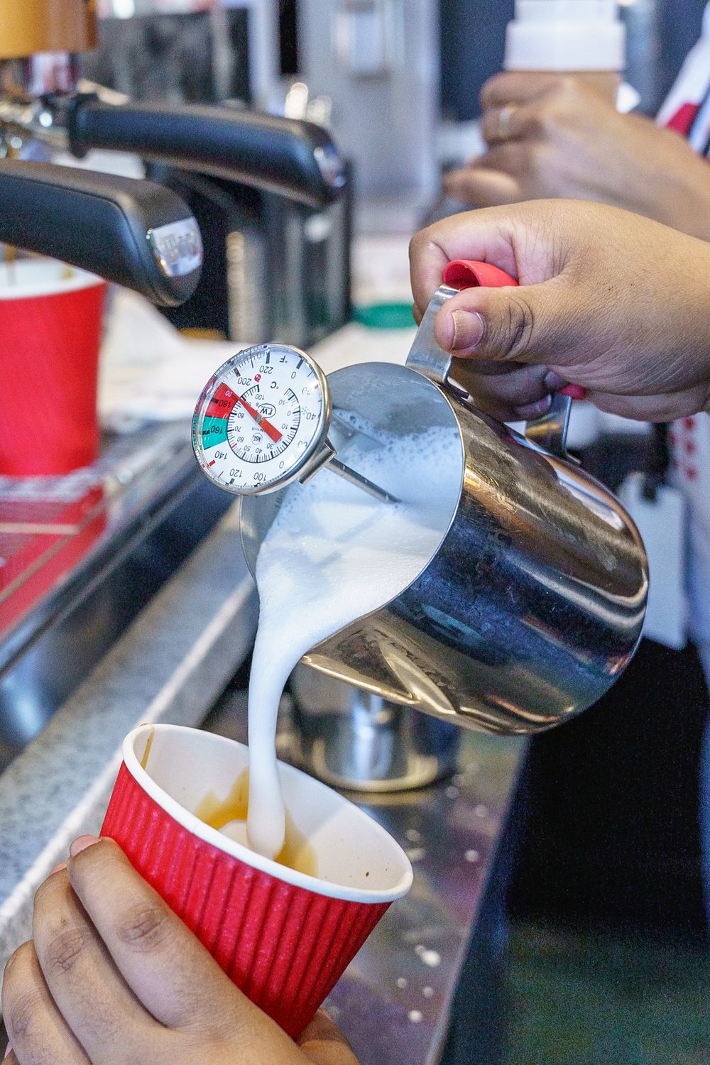 Steamed milk being poured from a metal pitcher into a red paper coffee cup.