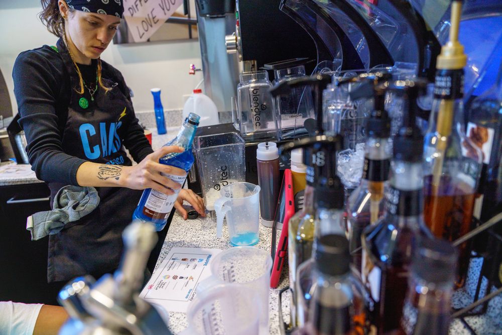Barista preparing drinks at a coffee shop counter with bottles and equipment.