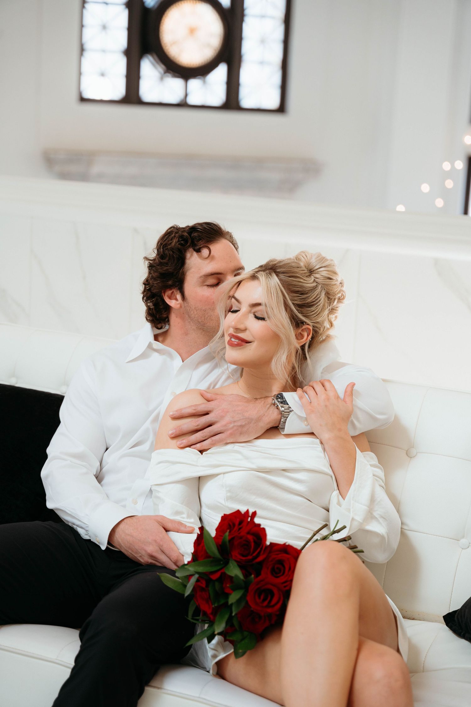 Romantic couple lounging on white couch with red roses creating intimate Valentine's Day atmosphere.