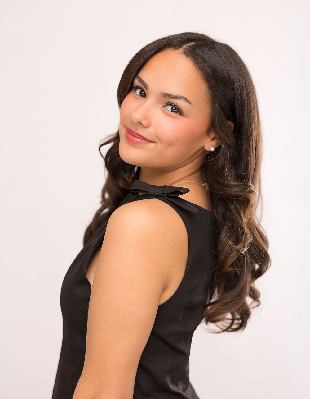Young woman with long dark wavy hair smiles over shoulder wearing black sleeveless dress against white background.