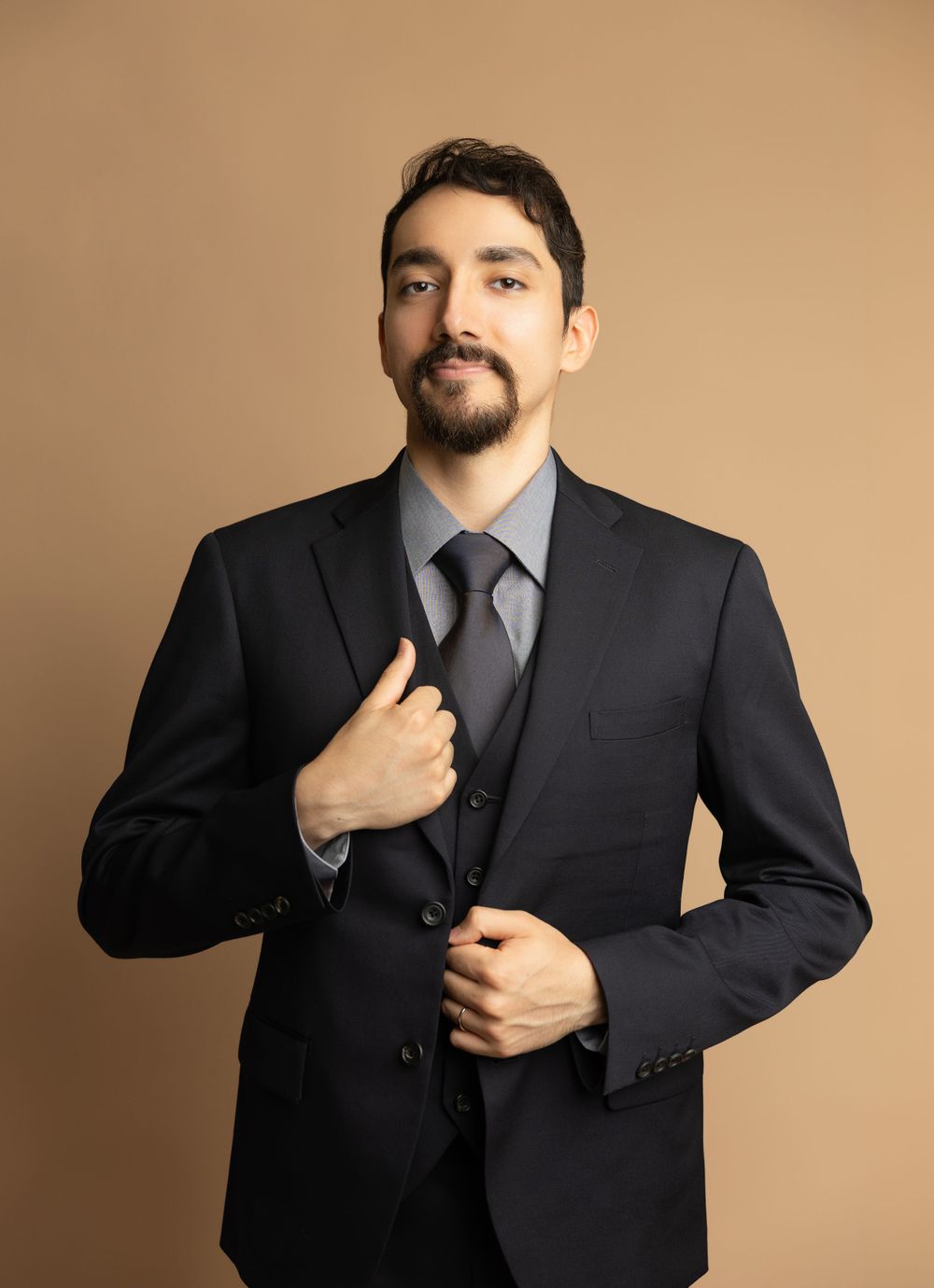 Professional portrait of person in dark three-piece suit adjusting tie against neutral background.