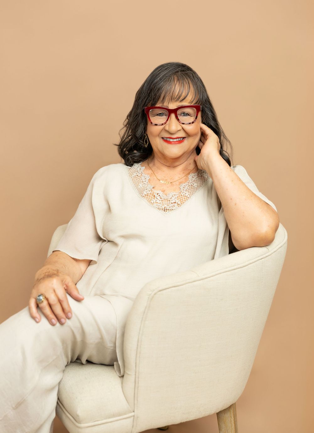 Person with glasses relaxing in cream chair wearing white outfit with statement necklace.