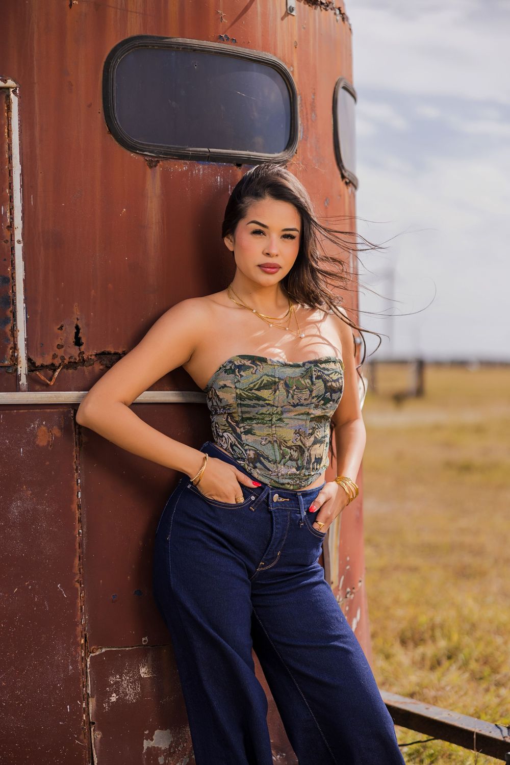 Young woman in strapless floral corset top and blue jeans leaning against rustic red trailer in open field.