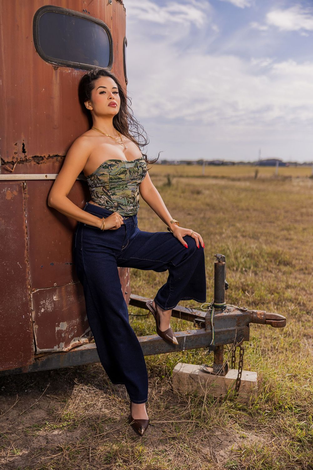 A woman in a camo corset and wide-leg jeans poses against a rusty vintage trailer in an open field.