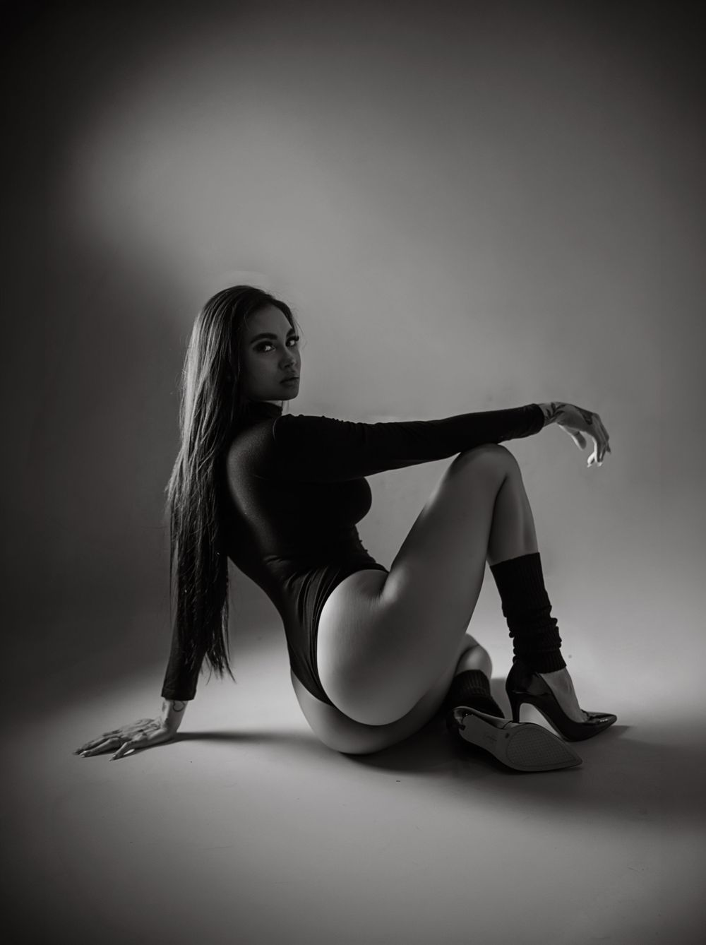A black and white image of a woman with long hair sitting in a provocative pose on a dark background.
