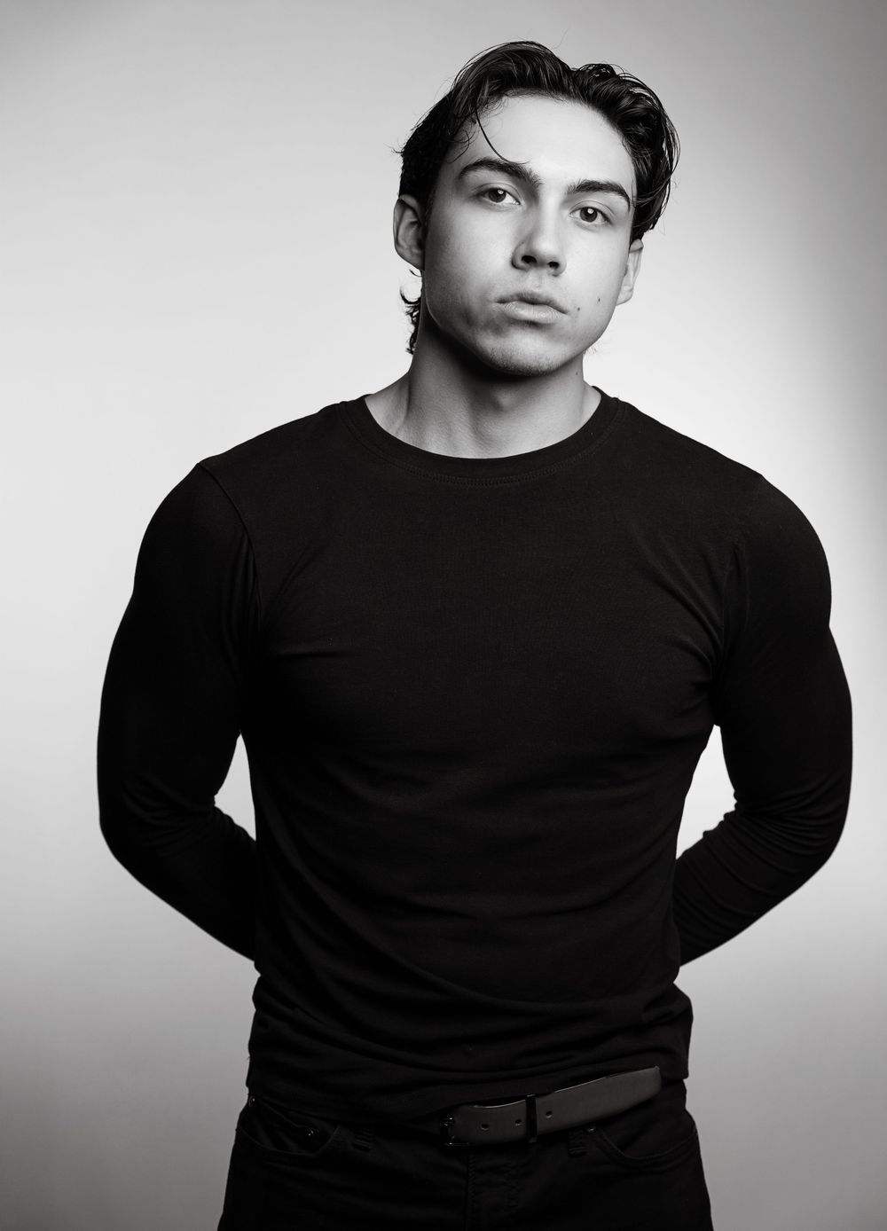 Black and white studio portrait of a young muscular man in a black long-sleeve shirt posing confidently.