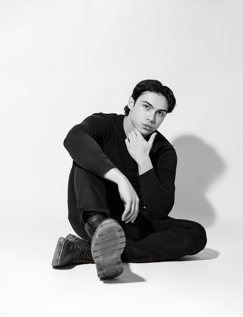Black and white photo of a young man in all-black outfit sitting cross-legged on white studio floor.