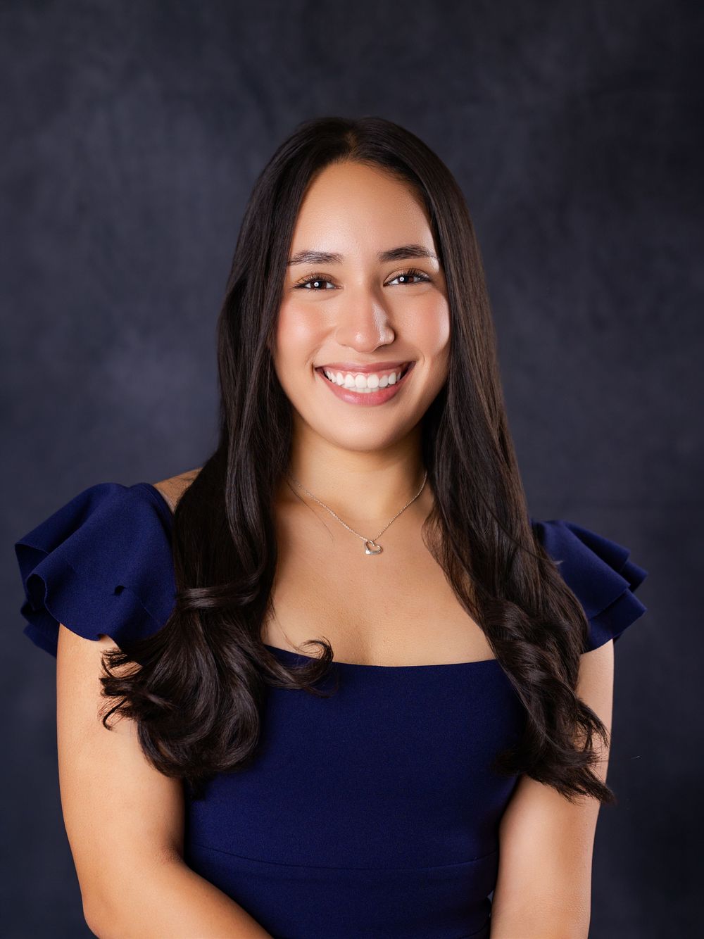 Young woman with long dark hair smiling in a navy blue dress, professional portrait with dark studio background.