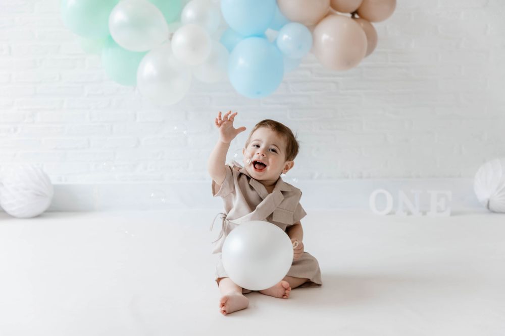 A young child playing with pastel balloons and soft toys on a white floor during a first birthday photoshoot.