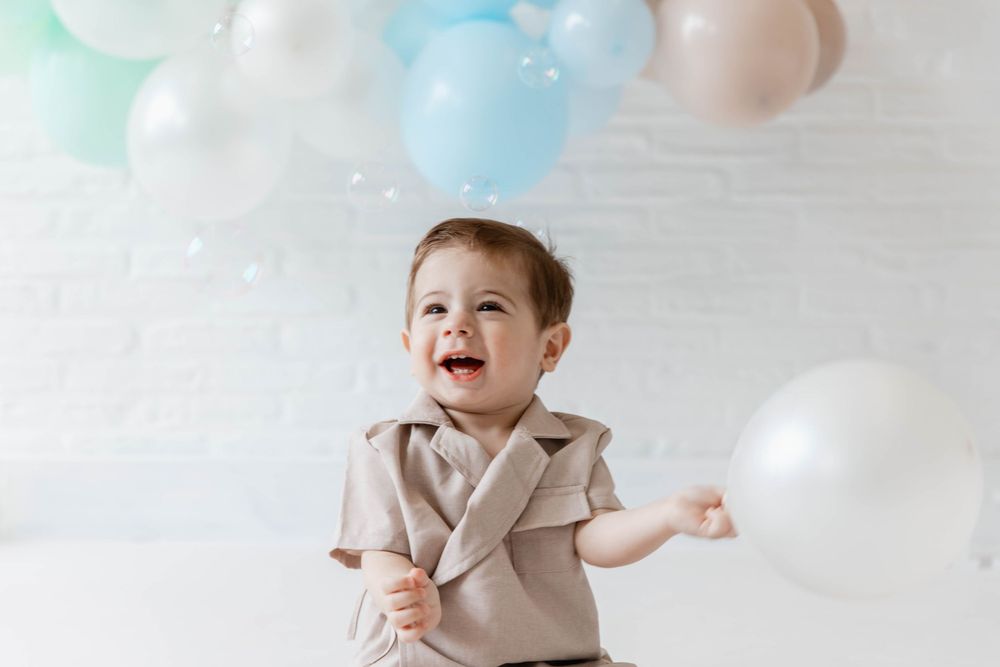 A happy small child in beige clothing smiles while sitting and playing among soft pastel colored balloons and clouds.