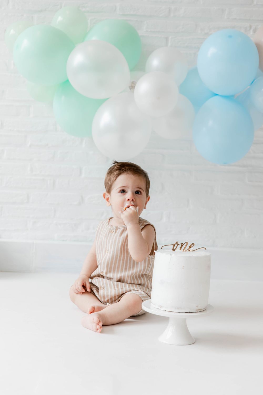 Simple white cake with 'one' topper and pastel mint and blue balloons for first birthday celebration against white backdrop.