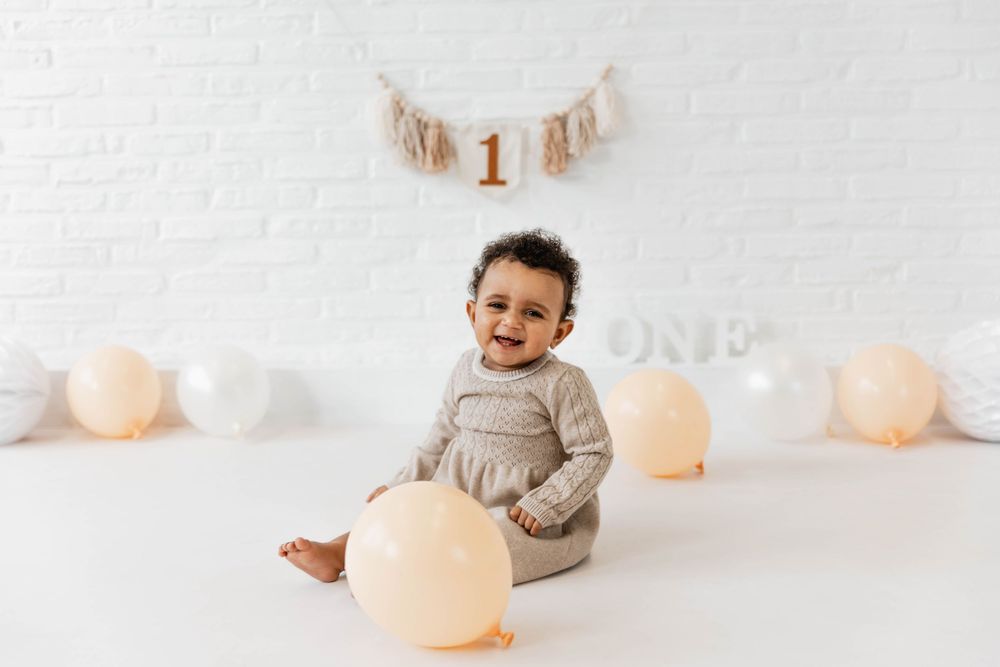 A baby in a gray sweater sits on white floor with peach and white balloons and a banner in the background.